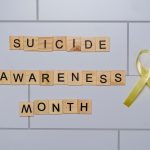 Blocos de madeira com a frase “Suicide Awareness Month” ao lado de uma fita amarela, símbolo de conscientização sobre a prevenção do suicídio.
