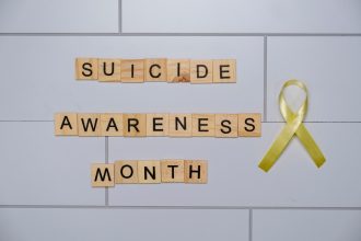 Blocos de madeira com a frase “Suicide Awareness Month” ao lado de uma fita amarela, símbolo de conscientização sobre a prevenção do suicídio.