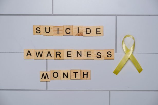 Blocos de madeira com a frase “Suicide Awareness Month” ao lado de uma fita amarela, símbolo de conscientização sobre a prevenção do suicídio.