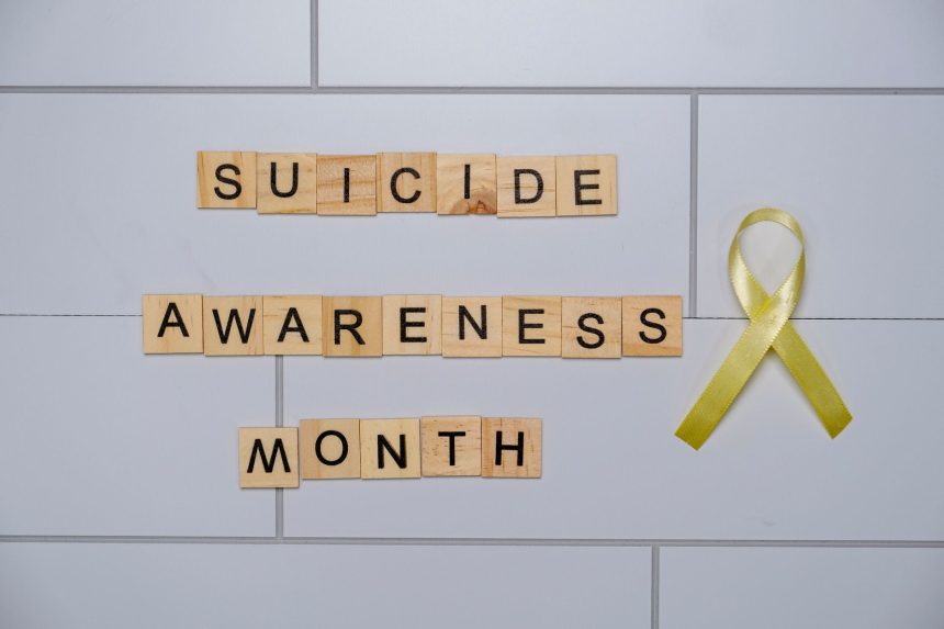 Blocos de madeira com a frase “Suicide Awareness Month” ao lado de uma fita amarela, símbolo de conscientização sobre a prevenção do suicídio.