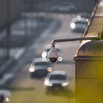 Câmera de monitoramento voltada para uma via urbana movimentada, com veículos desfocados ao fundo.