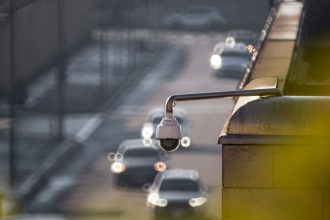 Câmera de monitoramento voltada para uma via urbana movimentada, com veículos desfocados ao fundo.