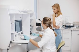 Duas profissionais de saúde analisam resultados de exames em um computador dentro de uma clínica.