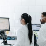 Dois pesquisadores de jaleco e óculos analisam dados em um computador dentro de um laboratório.