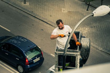 Trabalhador em uma plataforma elevatória realiza manutenção em um poste de iluminação pública em uma rua urbana movimentada.