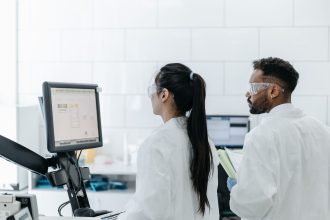 Dois pesquisadores de jaleco e óculos analisam dados em um computador dentro de um laboratório.