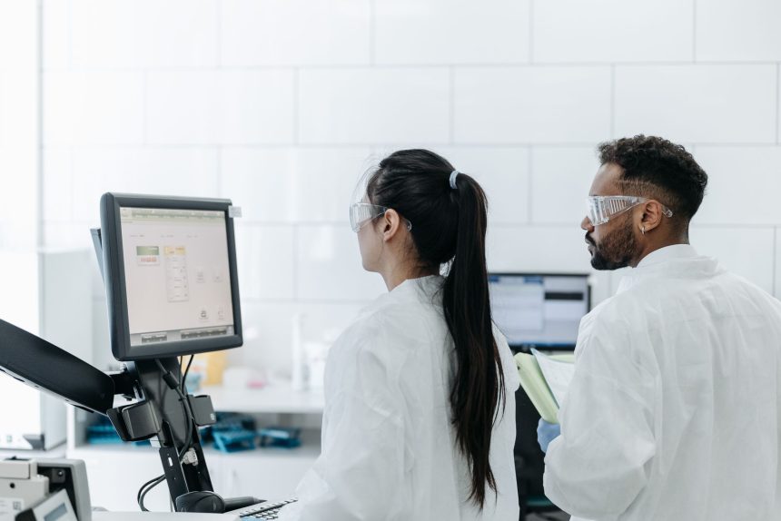 Dois pesquisadores de jaleco e óculos analisam dados em um computador dentro de um laboratório.